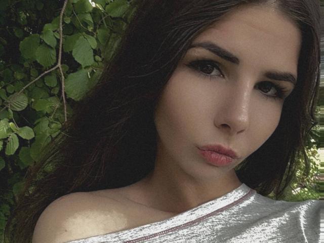 VickiLipoma has dark brown eyes, full pink lips, and long brown hair. She's wearing a white off-shoulder top, looking serious with a slight pout. Her skin is fair.