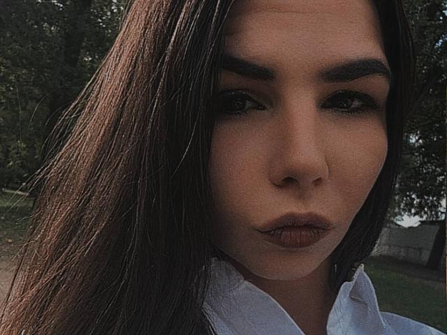 VickiLipoma a les cheveux bruns foncés et lisses qui lui couvrent un œil, des yeux marrons et des lèvres pleines et brunes. Elle porte une chemise blanche et a l'air sérieuse, avec un léger froncement de sourcils..