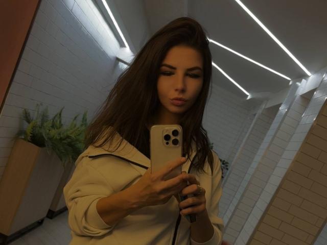 VickiLipoma with long brown hair, wearing a white jacket, taking a selfie in a dimly lit bathroom with white tile walls and ceiling lights, holding an iPhone.
