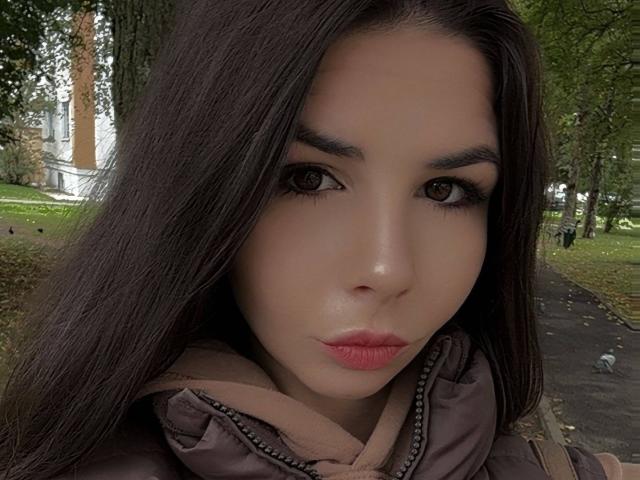 El rostro de VickiLipoma, ojos marrones, labios carnosos con lápiz labial rosa, cabello largo y oscuro, vistiendo una chaqueta marrón, expresión seria, un ligero rubor rosado en las mejillas, al aire libre..