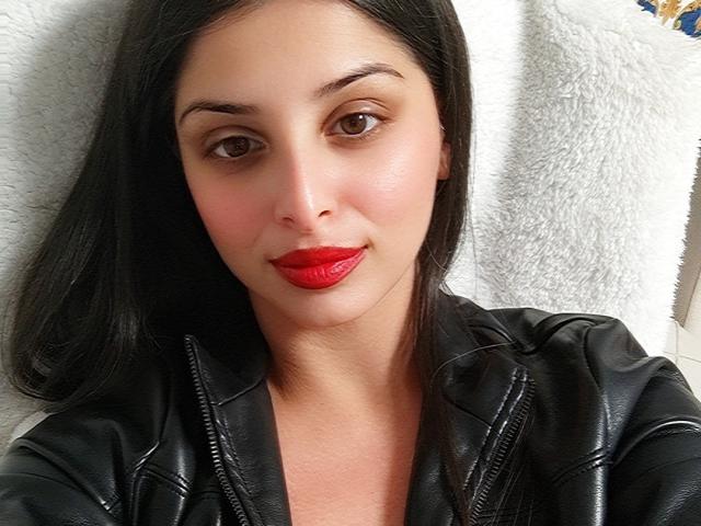 ScarletOra has dark brown eyes, red lipstick, black leather jacket, and straight black hair. Her face is slightly flushed. She's lying against a white, fluffy texture.