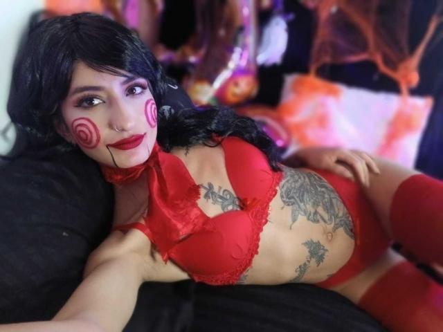 Kaiix with black hair, red lipstick, and red makeup dots, wearing red lingerie, tattoos on her stomach, lying on a black bed, red thigh-highs, and a red choker.