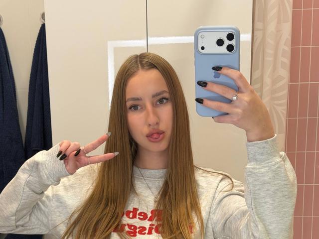 Dacia Schonhardt, con lunghi capelli biondi, una felpa grigia, smalto nero sulle unghie, la lingua fuori e che fa il segno della pace, sta scattando un selfie allo specchio con un iPhone blu..