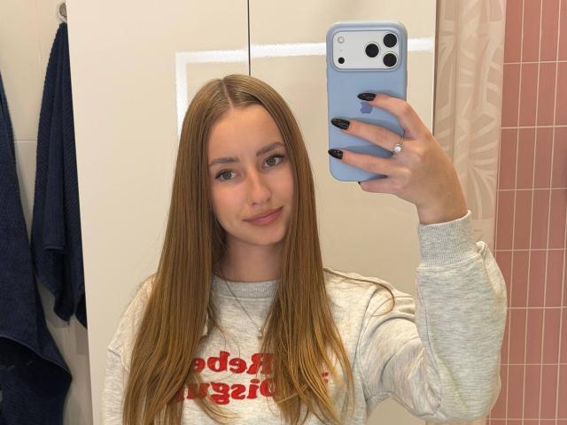 Dacia Schonhardt, met lang blond haar, een grijze blouse, zwarte nagels, die een blauwe iPhone vasthoudt, een spiegel-selfie maakt, lichtjes glimlachend, een ring draagt, in een badkamer..