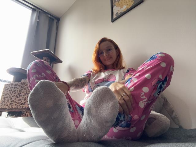 SarahRousen with red hair, glasses, pink polka dot pajamas, and grey socks, sitting with legs up, showing her bare feet and tattooed arm, on a grey couch.