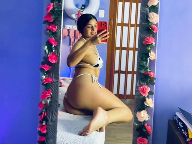 LonxEva in a white bikini, showing her ass and tits, kneeling on a bench, taking a mirror selfie with red phone, floral mirror frame, blue wall, wooden door.