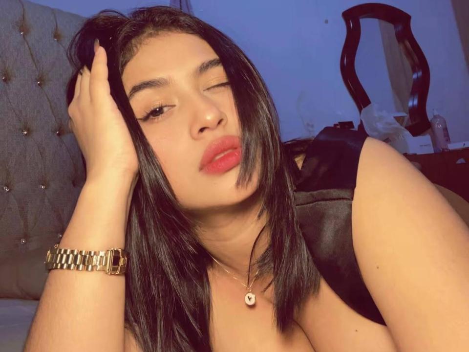Amaraluxx, avec ses cheveux noirs et son rouge à lèvres, fait un clin d'œil à la caméra, une main dans ses cheveux, portant une montre en or et un collier, dans une pièce faiblement éclairée, montrant ses seins..