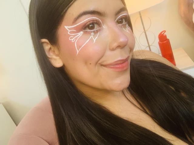 CatalinaReiyna with white face paint on her cheeks, smiling at the camera, long black hair, pink shirt, red bottle in the background, light skin, full lips, cute.