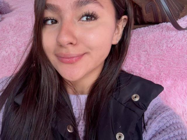 AlessiaFerry, avec de longs cheveux bruns, des yeux bruns et un petit sourire, portant un gilet noir par-dessus un pull violet, regardant l'appareil photo, allongée sur une couverture rose et douce..