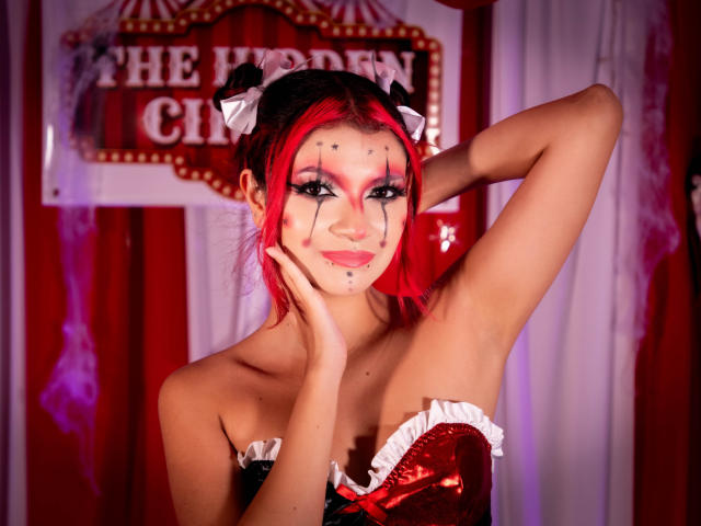 AriaRosel with red hair, black and white face paint, red and black corset, white frills, hand on cheek, arm raised, smiling, looking at camera, in a circus-themed setup.