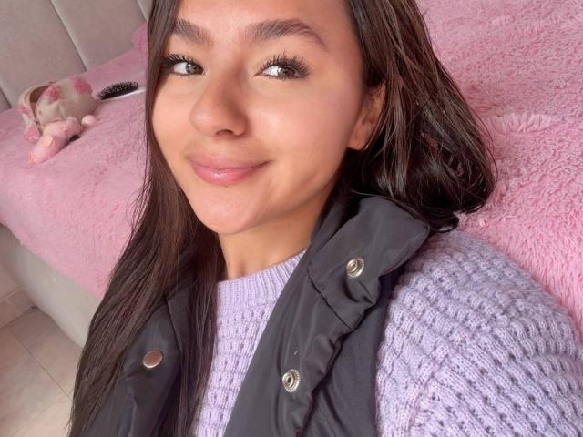 AlessiaFerry, avec ses longs cheveux bruns foncés, sa peau brun clair et son sourire charmant, porte un pull en tricot violet et un gilet noir, mettant en valeur ses petites seins galbés..
