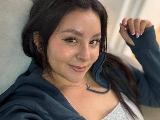 EmaJames, met donker haar, lichte huid, subtiele make-up, draagt een marineblauwe hoodie en een witte top, glimlacht licht, houdt haar arm omhoog, heeft roze nagellak, en draagt een ketting met een klein hangertje..