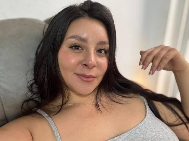 EmaJames has dark hair, a light skin tone, and is wearing a gray tank top. She's smiling with subtle makeup and a pink nail. Boobs are partially visible.