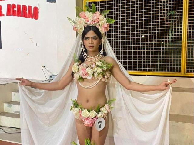 AudreyLustt est une femme à la peau foncée, avec de petites seins, portant des décorations florales, un voile blanc et une étiquette avec le numéro 1, debout, les bras tendus, dans un costume floral et nu..