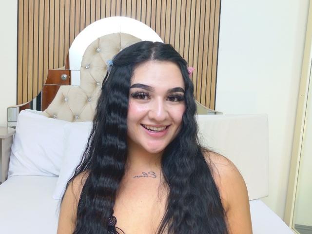 SamyBennet a de longs cheveux noirs ondulés, des sourcils foncés et un sourire qui montre ses dents. Elle porte un haut en dentelle noire et a un petit tatouage sur la poitrine..