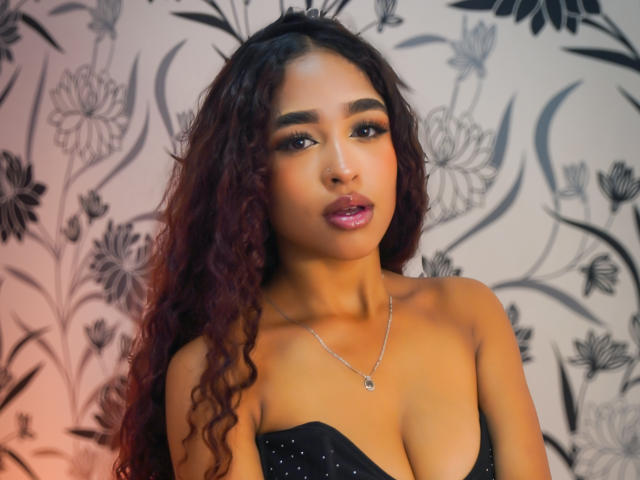 MadisonDawson has curly red-brown hair, dark eyes, glossy pink lips, and wears a black strapless top with small studs, showing her tits, and a silver necklace.