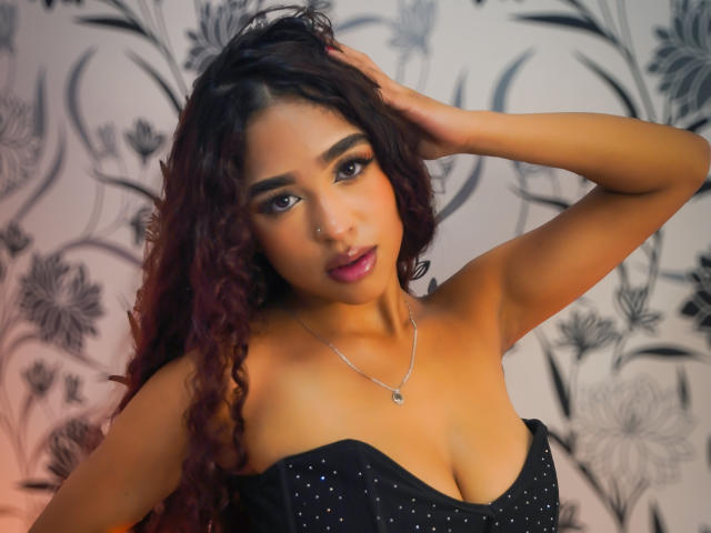 MadisonDawson with dark curly hair, medium brown skin, and full lips, wearing a black strapless dress with small sparkles, hand in hair, silver necklace, and a nose ring.
