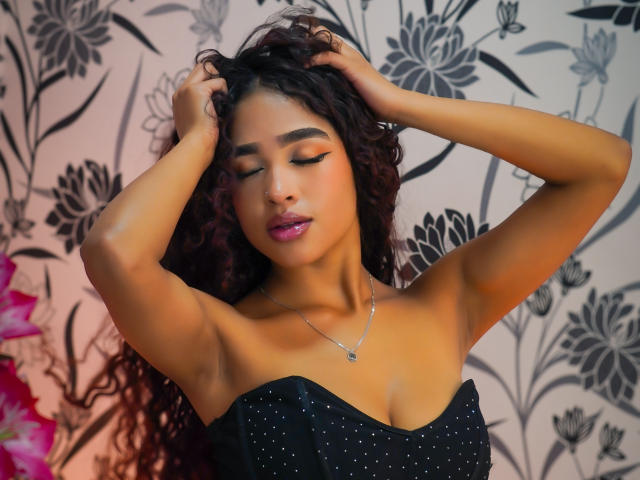 MadisonDawson with curly dark hair, hands in her hair, eyes closed, wearing a black strapless dress with sparkles, pink lipstick, and a silver necklace, against floral wallpaper.