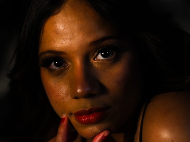 ZaraBlaze's dark brown eyes stare intensely, her glossy red lips slightly parted, with dark brown skin and long eyelashes. Her hand with red nails rests near her chin.