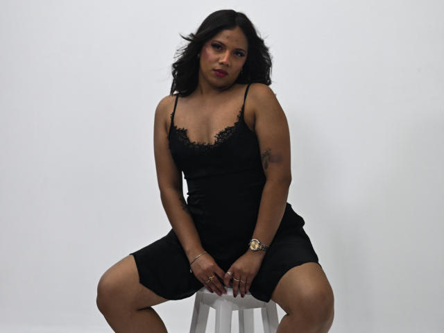 ZaraBlaze, a curvy Latina with dark hair, sits on a stool in a black lace-trimmed dress, legs spread, showing her ass, with a tattoo on her right arm.
