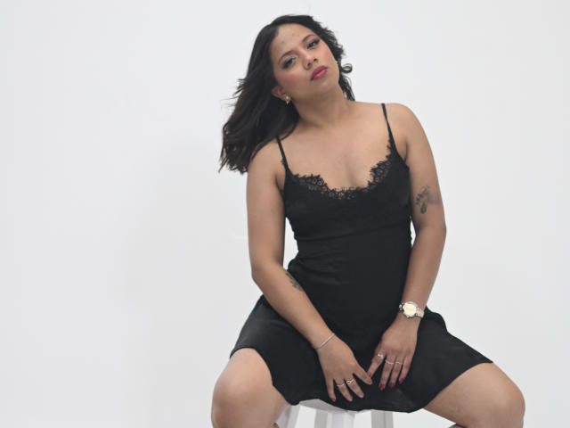 ZaraBlaze, a curvy Latina woman with black lace-trimmed black dress, sits on a stool with legs spread, showing her ass, tattooed arm, and red lipstick, wearing a gold watch.