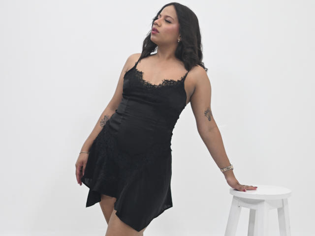 ZaraBlaze, vêtue d'une robe noire bordée de dentelle, se tient debout, une main posée sur un tabouret blanc, montrant ses tatouages sur les bras, les yeux fermés et les lèvres légèrement entrouvertes..