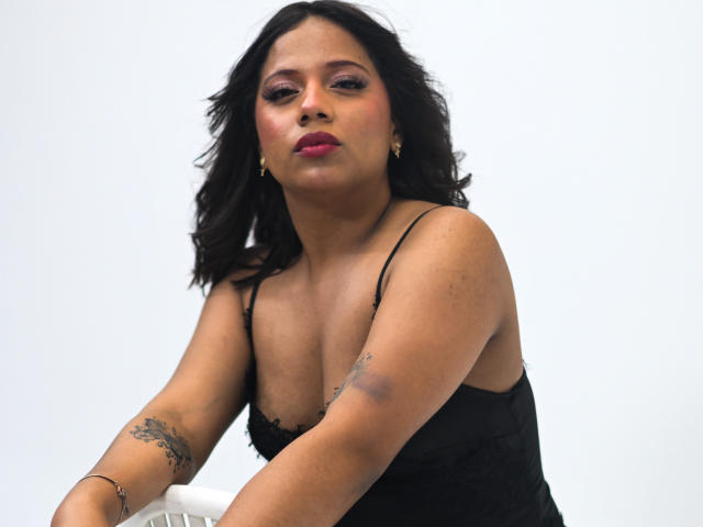 ZaraBlaze with medium brown skin, black wavy hair, pink lipstick, black tank top, tattoos on arms, puffy pink eyelids, gold earrings, puckering lips, white background.