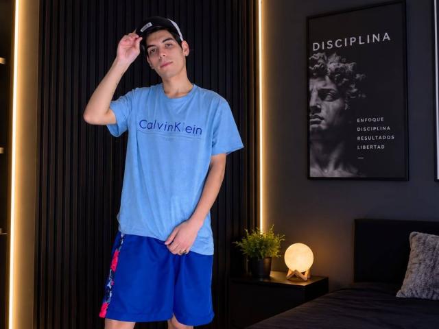 JaxMaxxi portant une chemise bleue Calvin Klein, un short bleu avec des rayures rouges et bleues, une casquette noire, debout près d'un lit avec une lampe et une plante en pot, regardant la caméra..