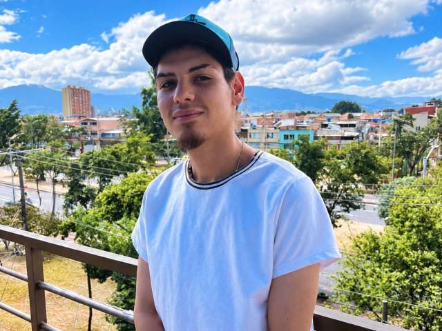 JaxMaxxi, com barba, usando um boné azul e camiseta branca, em pé em uma varanda, sorrindo levemente, com montanhas e céu nublado ao fundo..