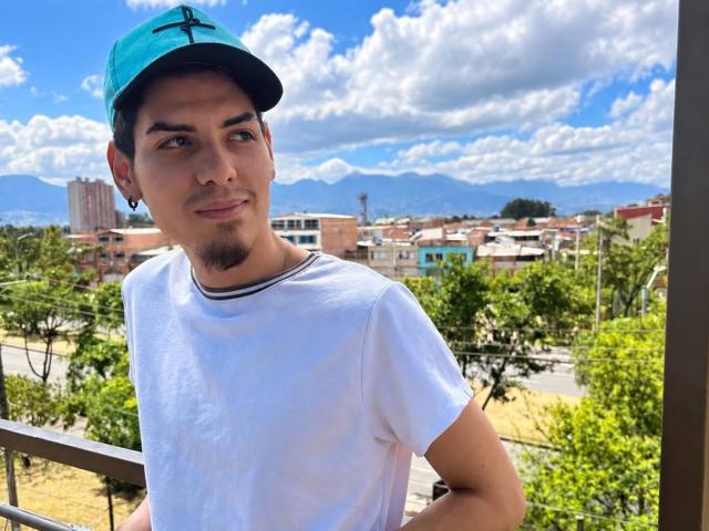 JaxMaxxi, con un berretto verde, una maglietta bianca e un piccolo pizzetto, in piedi su un balcone, che guarda la telecamera, cielo blu con nuvole soffici sullo sfondo..