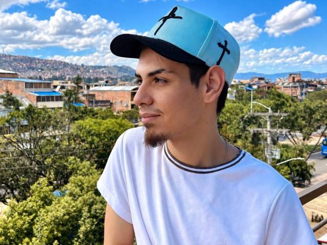 JaxMaxxi with a light blue cap, white tee, and goatee, looking sideways with a slight smile, short brown hair, and a stubble. Clear blue sky, fluffy clouds.