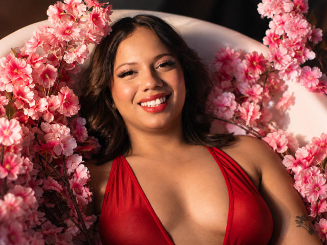 ZaraBlaze, com flores de cerejeira rosa ao seu redor, vestindo uma blusa vermelha, com decote profundo e transparente, sorrindo com batom vermelho, cabelo castanho ondulado e uma tatuagem no braço direito..