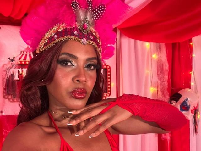 CamilaWest with red lipstick, dark makeup, and a pink feathered crown, wearing a red top and arm glove, pouting with hand near her face, showing her tits.