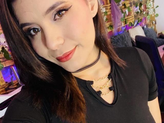LucieMilner with brown hair, red lipstick, black choker, gold necklace, black shirt, eyeliner, and a cute smile, looking at the camera. Her tits are small and she's pretty.