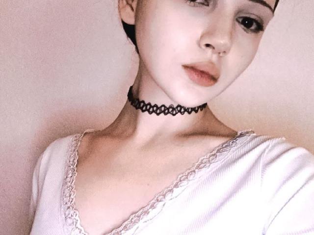 AlexasCherry, avec sa peau claire, porte un collier noir et un haut blanc avec des détails en dentelle. Elle a un petit anneau au nez et ses lèvres sont légèrement entrouvertes, et elle regarde doucement l'appareil photo..