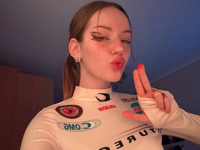JamesAughenbaugh with pouty lips, wearing a white racing suit, has long brown hair in a ponytail, winged eyeliner, nose ring, and pink lipstick, making a peace sign with red nails.
