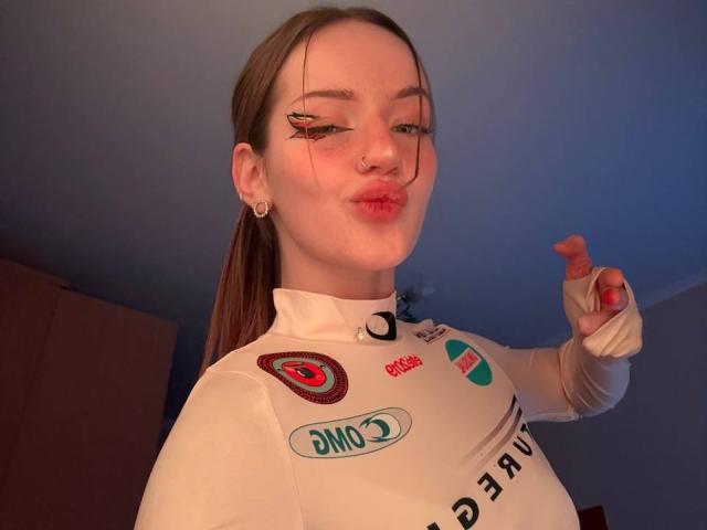 JamesAughenbaugh with pouty lips, wearing a white sports top, has long brown hair in a ponytail, nose ring, and black eyeliner. Pointing with a finger, showing a red nail.