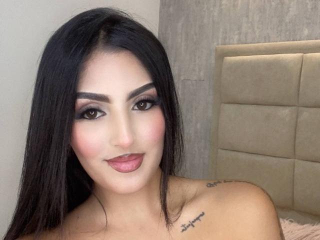 AriiaMon has long black hair, dark smoky eye makeup, pink blush, and pink lipstick. She's smiling with a tattoo on her right shoulder. She's topless.