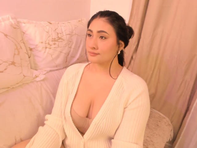 ElyRoss with dark hair in a bun, wearing a white ribbed cardigan, exposing her large, soft, pale boobs in a pink bra, sitting on a bed.