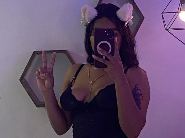 ChiaraBella with furry white cat ears, black lace bodysuit, and tattoo on right arm, taking a mirror selfie, showing off her tits and making a peace sign.