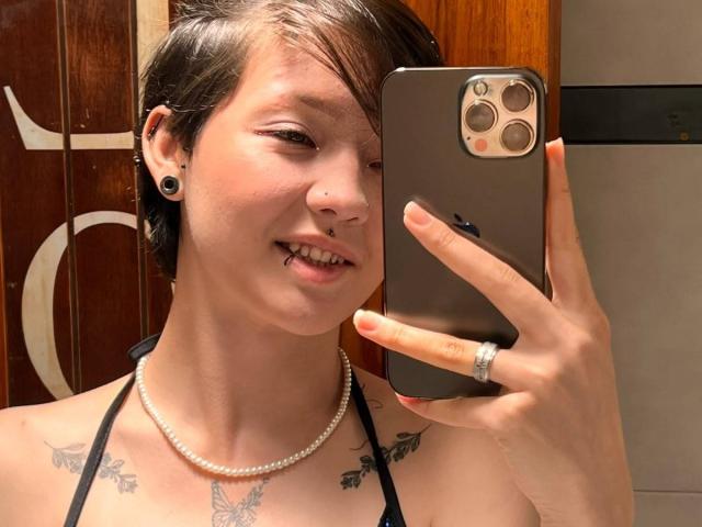 LissyLuxe with short brown hair, ear gauges, nose ring, tongue piercing, black halter top, pearl necklace, butterfly tattoos, holding a gold iPhone, smiling at the camera.