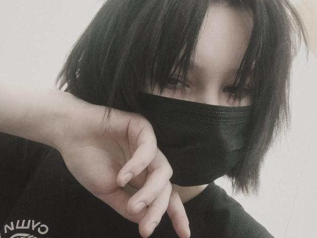ErikaStone con maschera nera, capelli neri corti, pelle pallida e camicia nera, che indica con il dito indice, occhi parzialmente visibili, watermark Nuiyo nell'angolo in basso a sinistra..