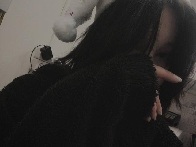 Erika Stone, con cabello oscuro, vestida con una bata negra y suave, cubriéndose la boca con la mano. Un peluche blanco con una marca roja en la cabeza. Habitación con poca luz..