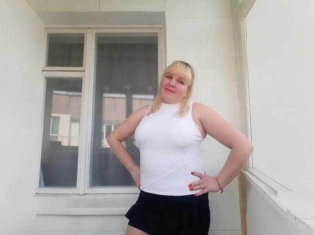 Leylisse mit blonden Haaren, einem weißen Top ohne Ärmel, schwarzen Shorts, mit den Händen an den Hüften, roten Nägeln, lächelnd, stehend vor einem Fenster, weiße Wände, leicht mollig, mittelgroße Brüste..