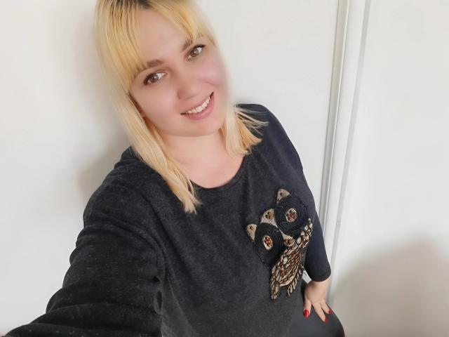 Leylisse, com cabelo loiro, sorrindo, vestindo um moletom cinza escuro com bordado de coruja, mão direita com esmalte vermelho, tirando uma selfie, mostrando seu rosto fofo e seios..