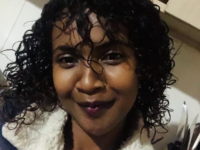 AaryaS has dark curly hair, brown skin, and a smile showing purple lipstick. She wears a fluffy white collar and looks slightly to the right.