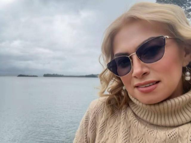 MisseScarleth, con i capelli biondi, indossa un maglione beige a collo alto e occhiali da sole scuri, ed è in piedi accanto a una superficie d'acqua calma, di colore grigio-blu. Ha la bocca leggermente aperta e un sorriso..