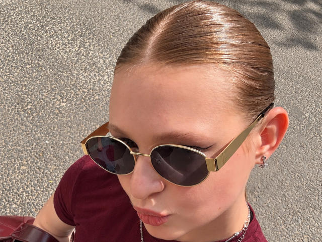 AkilahGhil has shiny brown hair pulled back, wearing gold-framed aviator sunglasses, a red shirt, and a small hoop earring in her left ear. Her nose is pierced.