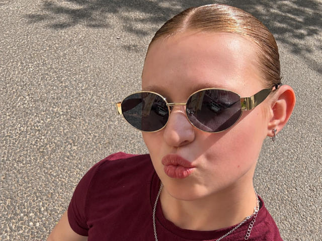 AkilahGhil with slicked-back brown hair, wearing dark aviator sunglasses, gold-tinted edges, pouty red lips, maroon t-shirt, and small hoop earrings, standing on a sunlit asphalt road.