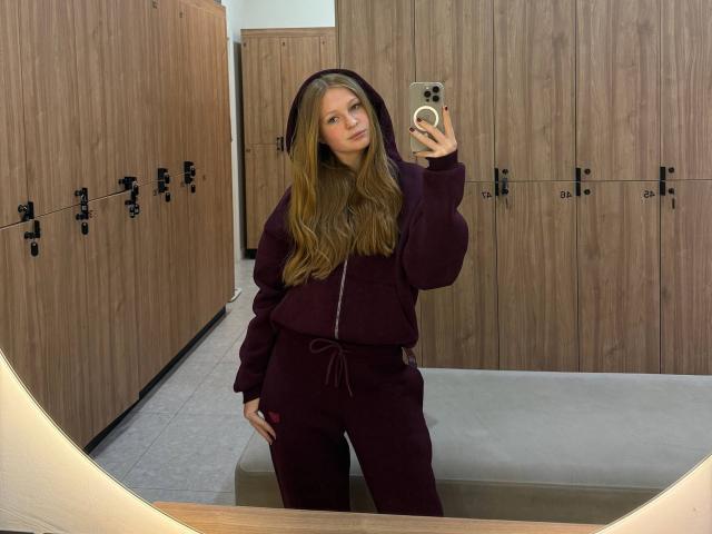 AkilahGhil with long brown hair, wearing a maroon hoodie and pants, taking a mirror selfie in a locker room. She's holding a gold phone, looking slightly bored.